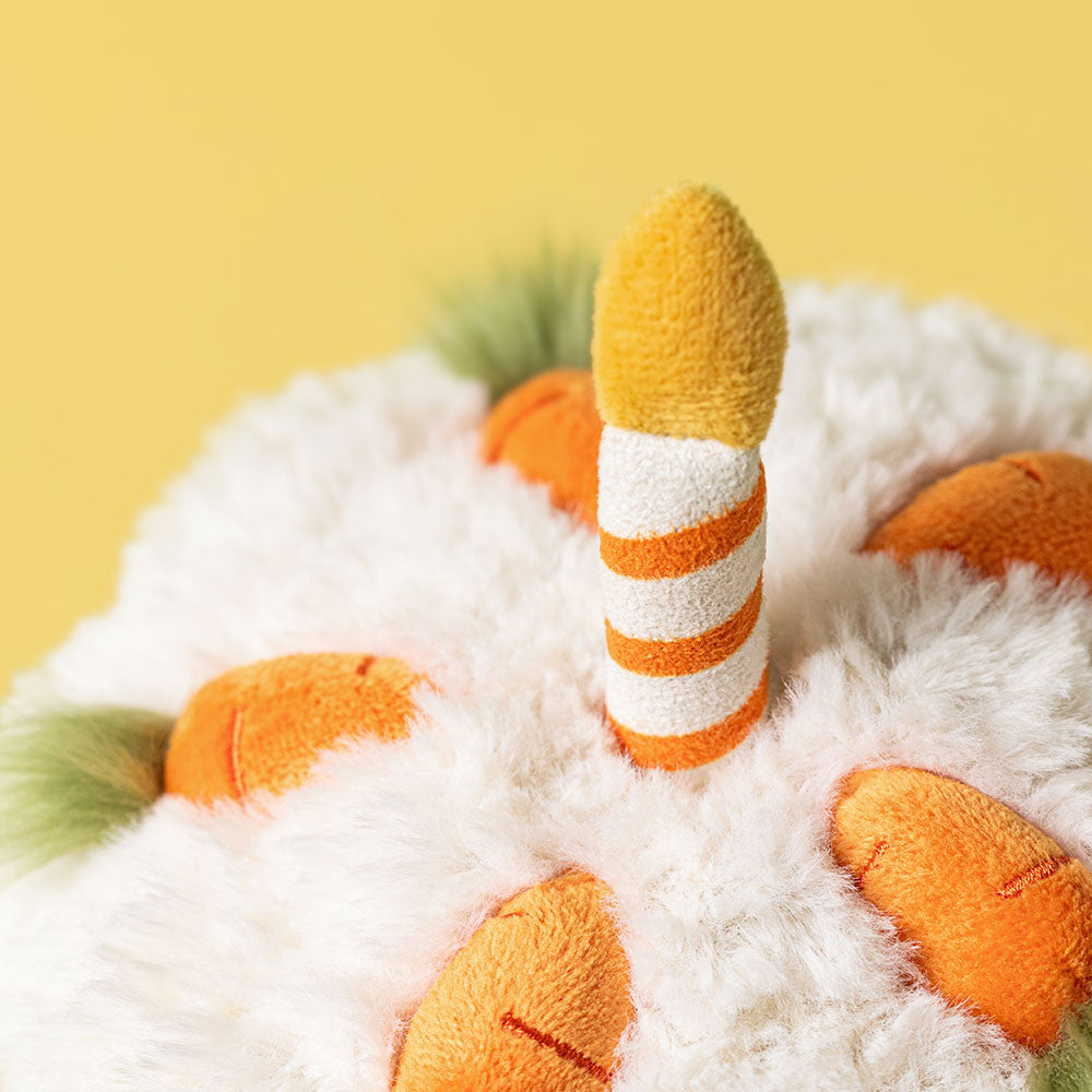 Die Oberseite ist mit weißem Plüsch, orangenen Stoff-Möhren und einer Kerze aus Velours verziert, Detailaufnahme vom jellycat Karottenkuchen