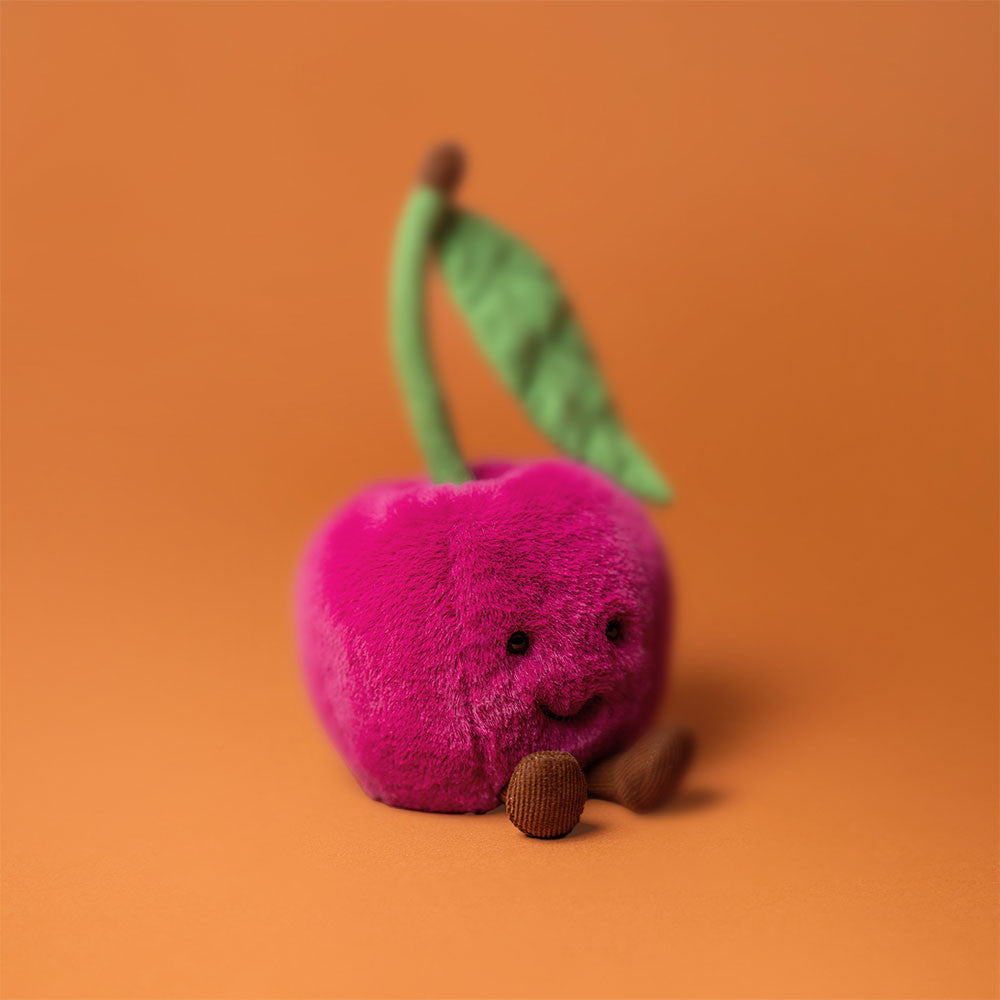 Flauschiger Jellycat Amuseables Cherry als Kirschen Plüschtier in Rot mit lächelndem Gesicht und weicher Oberfläche auf orangem Hintergrund, schräge Vorderansicht